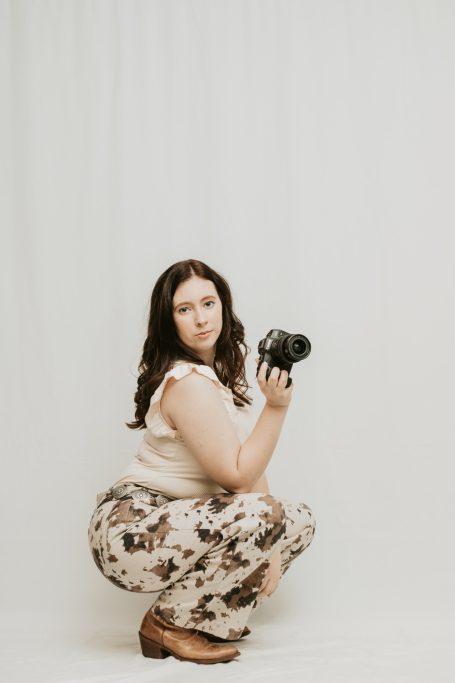Woman squatting with a camera, wearing a floral outfit and looking at the viewer.