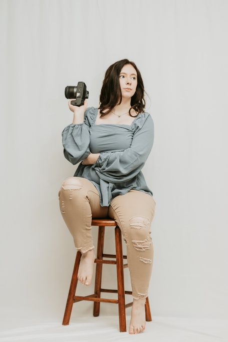 Woman sitting on a stool, holding a camera, wearing a gray top and beige pants.